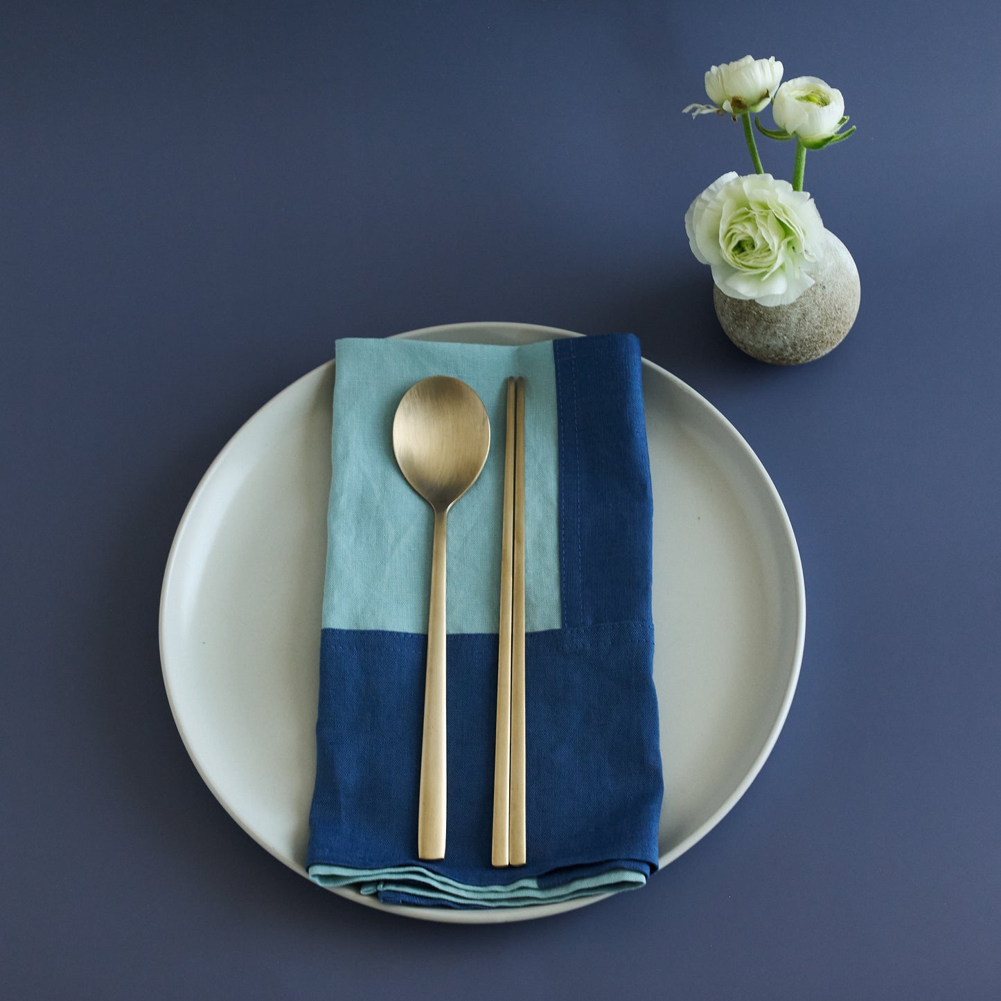 Dinner setting with a white plate, blue and green napkin, gold spoon, and chopsticks on a dark blue background.