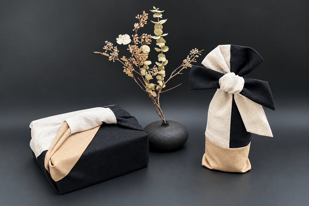 Two gift-wrapped packages on a dark studio background: one rectangular box wrapped in black paper with a beige and white fabric bow, and one cylindrical bottle wrapped in black and beige fabric with a large bow. A small vase with dried eucalyptus stands between them.